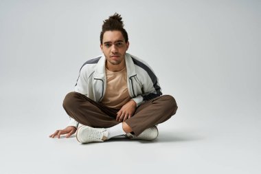 Genç bir Afrikalı Amerikalı, gri bir arka planda bağdaş kurmuş beyaz bir ceket, kahverengi pantolon ve beyaz spor ayakkabılar giyiyor..