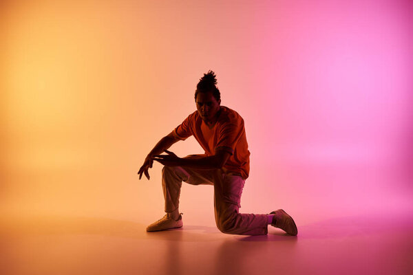 A young African American man in a vibrant orange shirt dances in a studio, illuminated by a gradient of pink and yellow lights.