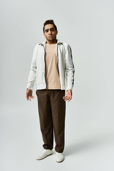 A young African American man in a white jacket, brown pants, and white sneakers poses against a grey background.