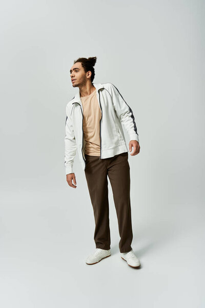 A young African American man poses in a stylish white jacket and brown pants against a gray background.