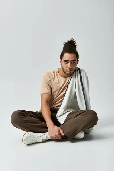 stock image A young African American man in a stylish outfit sits cross-legged on a grey background, posing confidently.