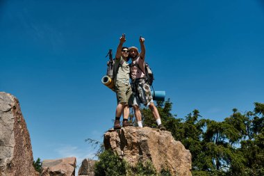 Two young men hike through summer wilderness, standing on a mountain peak. clipart