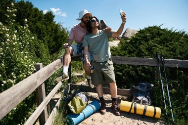 A young gay couple takes a selfie while hiking in the summer wilderness, enjoying the beautiful scenery and each others company. clipart