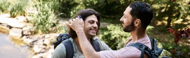 A young gay couple smiles and enjoys their hike through lush greenery on a sunny day. clipart