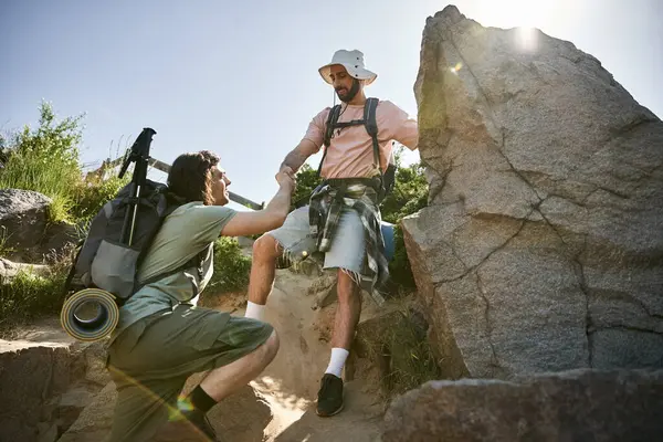 stock image A young gay couple hikes together in the wilderness on a sunny summer day, one man helping the other up a rocky path.