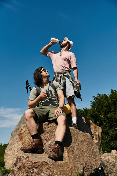 Two young men, a gay couple, hike and take a break on a rocky outcrop during a sunny summer day.