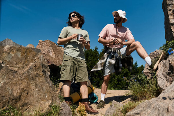 Two young men, dressed in casual summer attire, stand together on a rocky hiking trail, enjoying the view.