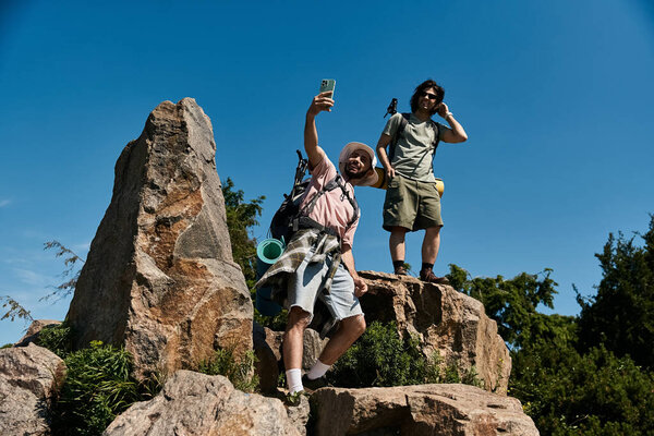 Two young gay men hike and take a selfie on a rocky mountaintop during a summer day.