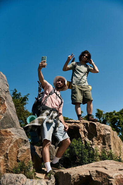 Two young men hike together in the wilderness, taking a selfie on a rocky mountaintop.
