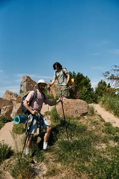 stock image Two young men are hiking together in a rocky landscape. They are both wearing backpacks and carrying hiking poles.