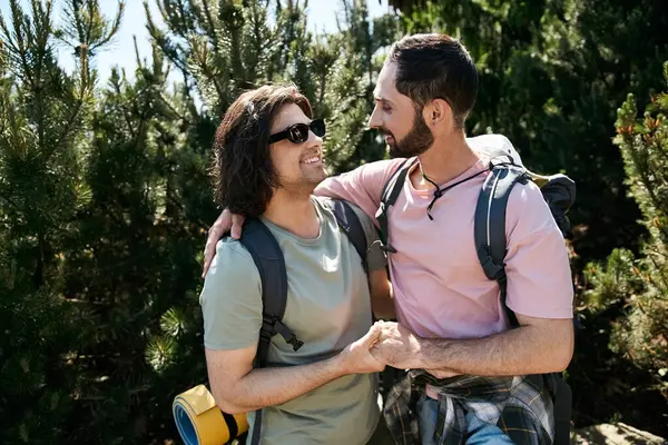 stock image Two young men, a gay couple, hike through a sunny summer forest, smiling and holding hands.