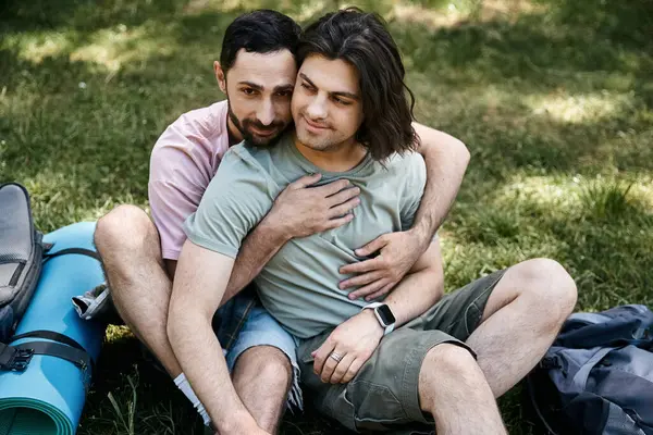 stock image Two young men, a gay couple, embrace while hiking in a lush green forest during the summer. Sitting on a mat with nearby backpacks.