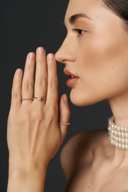 Yüzüklerle süslenmiş bir kadın eli, yüzüne yaslanmış, narin bir inci kolye sergiliyor..