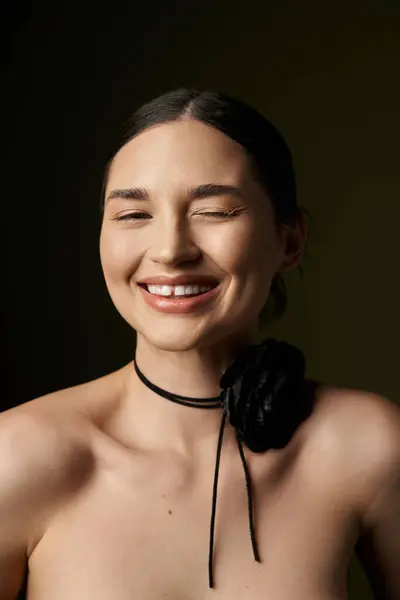 stock image A young woman with brunette hair smiles while wearing a black flower choker against a dark background.