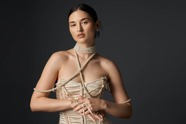 Молодая женщина с темными волосами и жемчужным ожерельем позирует драматично на темном фоне.