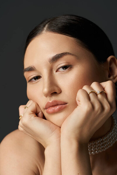 Молодая женщина с темными волосами и безупречной кожей кладет подбородок на руки. Она носит золотые кольца.