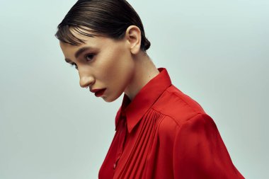 Kırmızı bluzlu şık bir genç kadın, gri bir arka plan önünde kendine güvenle poz veriyor, şık tavrını sergiliyor..