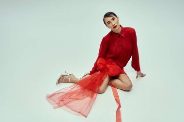 Kısa saçlı, kırmızı elbiseli, fiyonklu genç bir kadın gri bir arkaplanda poz veriyor..