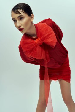 Kısa siyah saçlı genç bir kadın gri bir fonda kırmızı bir elbise içinde poz veriyor..