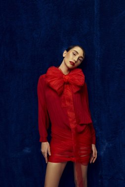 Kısa saçlı genç bir kadın, kırmızı elbiseli, büyük bir yayla mavi bir arkaplanda poz veriyor..