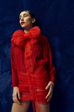 Kısa saçlı güzel bir kadın kırmızı elbiseyle poz veriyor. Mavi arka planda büyük bir fiyonk var..