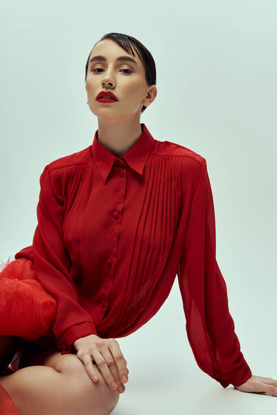 Красивая молодая женщина с короткими темными волосами позирует в красной рубашке на сером фоне.