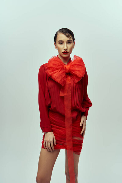 Молодая женщина с короткими темными волосами позирует в красном платье с большим бантом. Она стоит перед серым фоном.