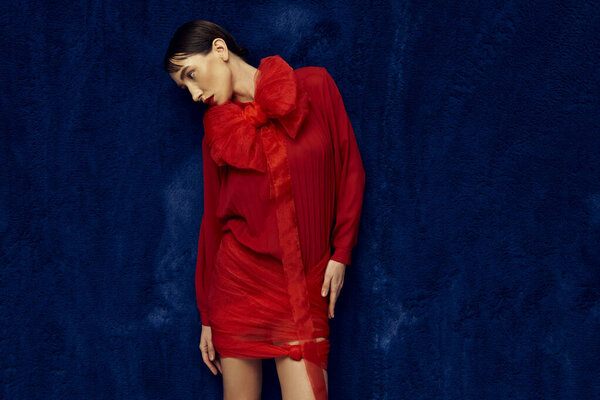 Молодая женщина в красном платье с бантиком позирует на синем фоне.