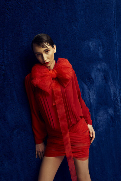 Молодая женщина с короткими волосами позирует в красном платье с большим бантом на синем фоне.