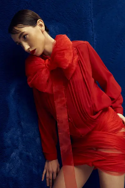 Stock image A young woman with short dark hair poses in a red dress with a large bow against a blue background.