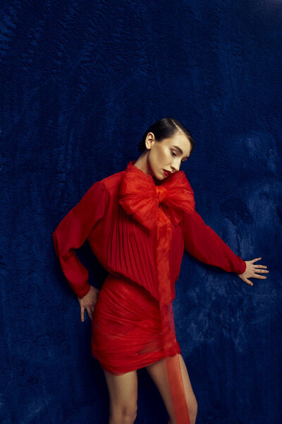 A young woman in a red dress with a large bow poses against a blue backdrop.