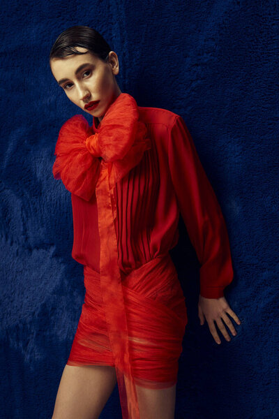 Молодая женщина с короткими волосами позирует на синем фоне, одетая в яркое красное платье с большим бантом.