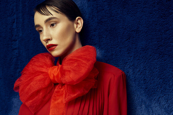 Молодая женщина с короткими волосами позирует в красном наряде с большим бантом на синем фоне.