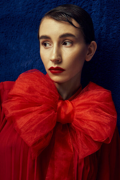 Молодая женщина с короткими волосами позирует в красной одежде с большим бантом, установленным на синем фоне.