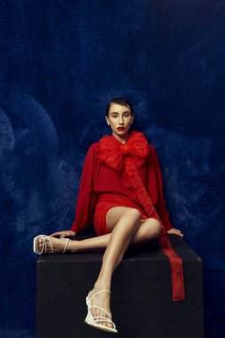 A young woman with short hair poses in a red outfit with a large bow against a blue background. clipart