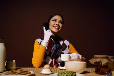 Şık giyinmiş genç bir kadın, elinde bir fincan kahve ve klasik bir telefonla bir masada oturuyor..