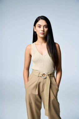 Genç bir kadın, krem rengi atlet bluz ve kahverengi pantolon giyen, kendine güvenen bir şekilde poz veriyor..