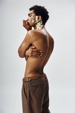 Genç, kaslı Afrikalı Amerikalı bir adam gri bir arka plana karşı poz veriyor, yüzü ve vücudu narin çiçeklerle süslenmiş..