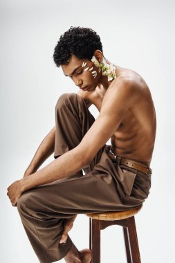 Genç, kaslı bir Afrikalı Amerikalı adam gri bir arka plana karşı yüzünde ve vücudunda beyaz çiçeklerle bir taburede oturuyor..