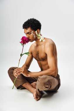 Çiçek kaplı Afro-Amerikan bir adam oturur, bacakları çapraz, nazikçe pembe bir gülü koklar.