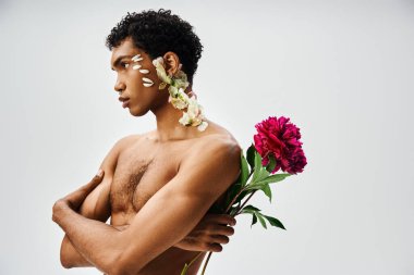 Genç, kaslı Afrikalı Amerikalı bir adam gri bir arka plana karşı poz veriyor, yüzünde ve vücudunda çiçekler var..