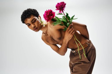 Genç, kaslı Afrikalı Amerikalı bir adam gri bir arka plana karşı poz veriyor, vücudunu ve yüzünü süsleyen çiçeklerle..