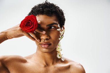Genç, kaslı Afrikalı Amerikalı bir adam gri bir arka plana karşı poz veriyor, yüzü ve vücudu çiçeklerle süslenmiş..