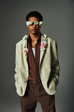 Genç bir Afrikalı Amerikalı şık bir ceketle poz veriyor. Gri bir arkaplanda yüzünü ve boynunu süsleyen çiçekler var..