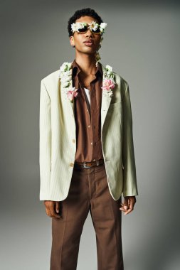 Genç Afrikalı Amerikalı bir adam şık bir ceketle poz veriyor, boynundaki ve yüzündeki çiçeklerle süslenmiş, gri bir arka planda..