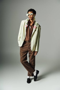 Genç bir Afrikalı Amerikalı, gri bir arka planda şık bir ceket giyen, boynunu ve yüzünü süsleyen çiçeklerle poz veriyor..