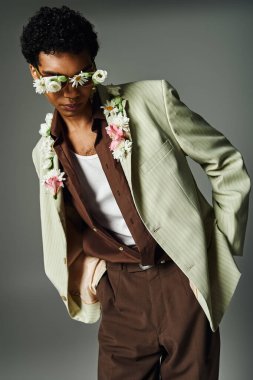 Genç Afrikalı Amerikalı bir adam şık bir ceket giyiyor ve çiçek aksanıyla gri bir arkaplanda poz veriyor..