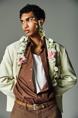 Genç bir Afrikalı Amerikalı şık bir ceketle poz veriyor, boynunda ve yüzünde gri bir arka planda çiçekler var..