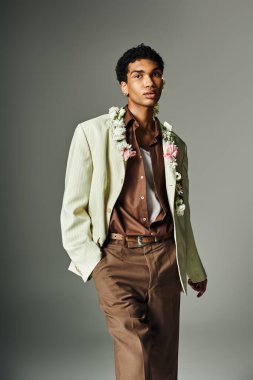 Genç bir siyah adam gri bir arka planın önünde duruyor açık yeşil bir ceket, kahverengi gömlek ve çiçek çelengi giyiyor..