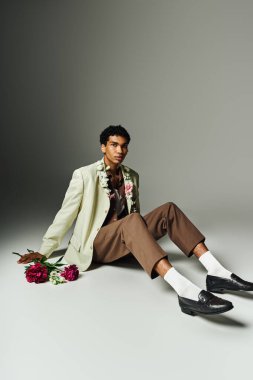Genç Afrikalı Amerikalı bir adam gri bir arka planda oturuyor, şık bir ceket ve çiçekli detaylar giyiyor.